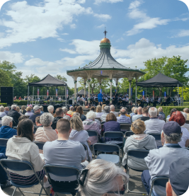 Lots of people sitting in a park looking at a bandstand on a summers day